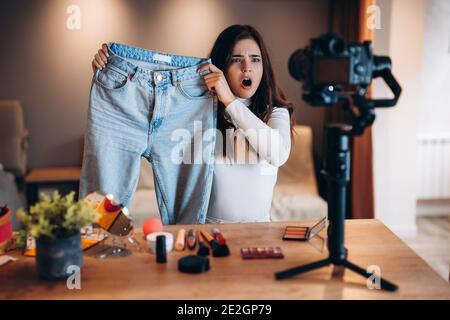 Beauty-Blogger mit dekorativer Kosmetik, die Jeans vor der Videokamera hält. Junge Frau ist überrascht und mit Streaming auf Kamera Stockfoto