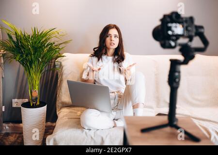 Junge, überraschte Bloggerin sitzt auf dem Sofa mit Laptop, der ihren Rede-Vlog vor ihrem Publikum aufzeichnet. Bloggen in Innenräumen Stockfoto