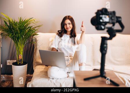 Junge, überraschte Bloggerin sitzt auf dem Sofa mit Laptop, der ihren Rede-Vlog vor ihrem Publikum aufzeichnet. Bloggen in Innenräumen. Live-Streaming Stockfoto