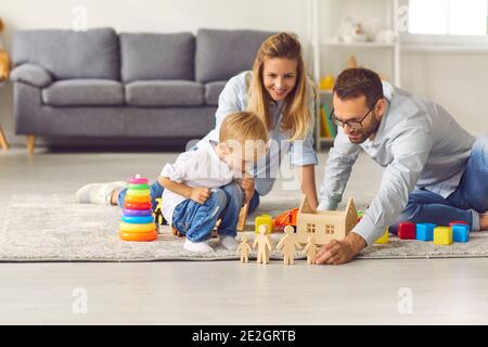 Junge Eltern spielen mit ihrem Sohn Holzfiguren, die auf dem Boden unter verschiedenen Spielzeugen sitzen. Stockfoto