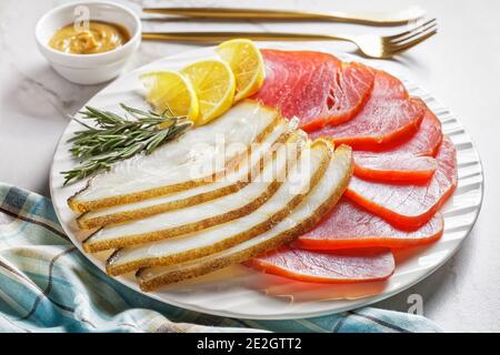 Geräucherter Fischteller: Kalter geräucherter Heilbutt und kalte geräucherte Thunfischscheiben auf einem weißen Teller mit Rosmarinzweigen, Zitronenkeilchen und englischem Senf Stockfoto