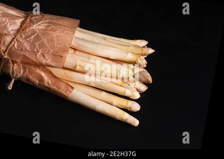 Nahaufnahme von weißem Spargel auf schwarzem Hintergrund.Weißer Spargel in Papier verpackt.frisches Gemüse mit einer Schnur auf schwarzem Hintergrund gebunden. Stockfoto