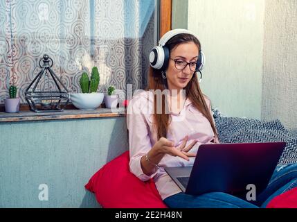 Schöne Frau, die von zu Hause aus auf einem roten Sofa sitzt Mit ihrem Laptop und mit Kopfhörern mit jemandem reden Stockfoto