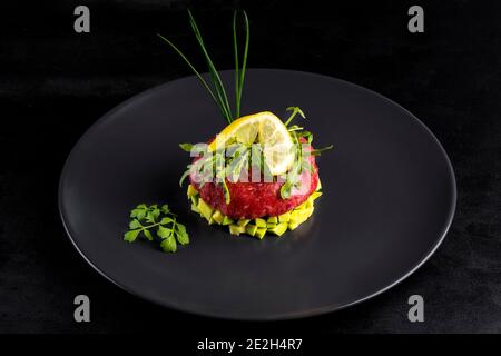 Rindertartar mit Ei auf einem schwarzen Steinteller serviert. Tartar auf schwarzem Teller mit Avocado, Kräutern, Salz und Hühnerdotter. Rohes Tartarfleisch.Schwarzer Backgro Stockfoto