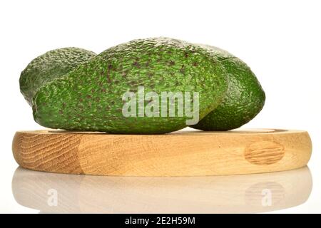 Drei grüne reife Avocados, einer im Vordergrund, im Fokus. Die beiden anderen sind im Hintergrund. Die Früchte liegen auf einem hölzernen runden Ständer, auf einem weißen Stockfoto