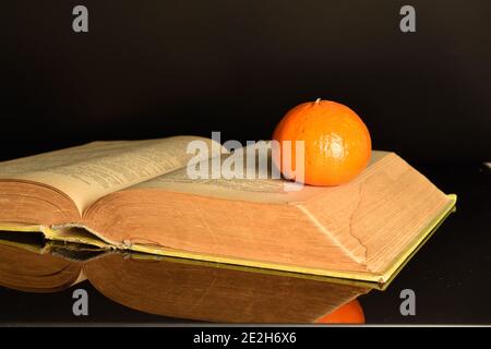 Auf einem offenen Buch auf schwarzem Grund liegt eine ganze runde, leuchtend orange saftige reife Mandarine. Stockfoto