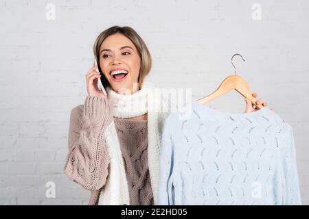 Fröhliche Frau in warmen Kleidern, die am Handy spricht und hält Kleiderbügel mit Pullover auf weißem Hintergrund Stockfoto