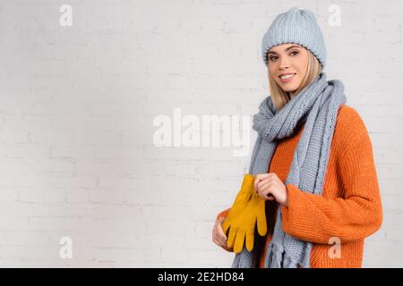 Fröhliche Frau in Strickmode hält Handschuhe in der Nähe von weißen Ziegel Wand Stockfoto