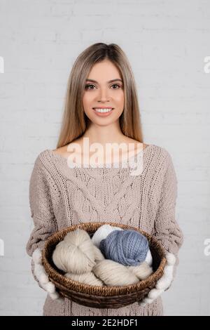 Junge Frau in Pullover und Handschuhe lächelnd, während sie den Korb hält Mit Strickfäden auf weißem Hintergrund Stockfoto