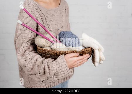 Beschnittene Ansicht der jungen Frau hält Korb mit Stricknadeln, Garn und Handschuhe auf weißem Hintergrund Stockfoto