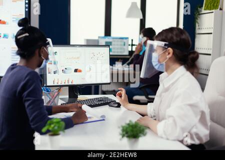 Afrikanischer Mitarbeiter, der mit Kollegen zusammenarbeitet, um Finanzgraph zu überprüfen und Gesichtsmaske für covid19 zu tragen. Multiethnisches Team arbeitet in Unternehmen mit neuen normalen Respekt soziale Distanz während der globalen Pandemie mit Coronavirus. Stockfoto