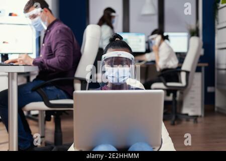 Afrikanischer Freiberufler am Arbeitsplatz mit Laptop trägt Gesichtsmaske gegen covid19. Multiethnisches Geschäftsteam, das während der globalen Pandemie mit Coronavirus soziale Distanz respektiert. Stockfoto