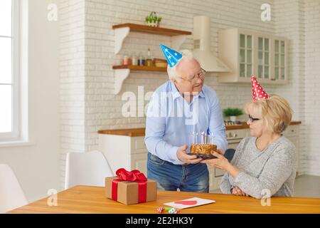 Romantischer älterer Mann gibt seiner Frau einen Kuchen, um einen Wunsch zu machen und die Kerzen auszublasen. Senior paar in farbigen Kegelhüte Geburtstag feiern oder Stockfoto