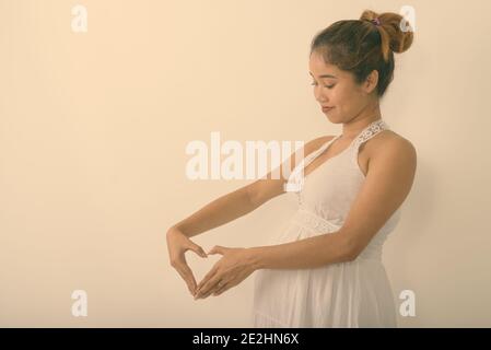 Profil anzeigen von jungen asiatischen schwangere Frau, Herz Hand neben ihrem Magen vor weißem Hintergrund Stockfoto