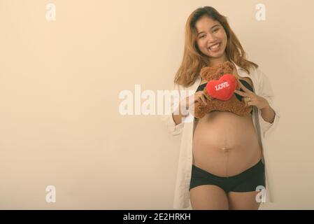 Studio shot von jungen asiatischen schwangere Frau glücklich lächelnd, während sie Teddybär mit Herz und Liebe Zeichen gegen weißen Hintergrund Stockfoto