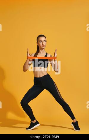 Sportlich schöne Frau in schwarzer Sportbekleidung Training mit Gummiband isoliert auf gelbem Hintergrund. Gesunder Lebensstil Stockfoto