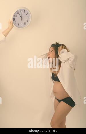 Profil anzeigen von jungen asiatischen schwangere Frau geschockt gleichzeitig Wanduhr mit weißem Hintergrund suchen Stockfoto