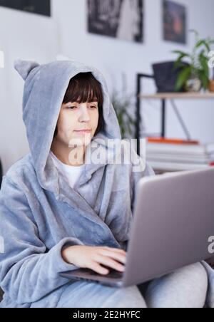 Brunette attraktives Mädchen in grauem Bademantel sitzt auf einem Bett und macht Abstand Job, Studium oder Online-Shopping über das Internet in einem Laptop. Remote-Arbeit c Stockfoto