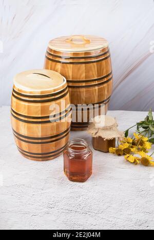 Zwei Keramikfässer und zwei Glasgefäße mit Honig und gelben Blüten auf hellem Hintergrund. Stockfoto