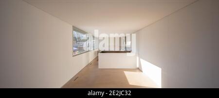 Großes und langes leeres Zimmer in einer Einfamilienwohnung. Riesige Fenster mit schöner Aussicht und der Himmel ist blau. Ein getrocknetes Licht dringt ein und trifft die Wand. Stockfoto