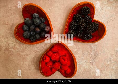 Verschiedene gesunde, frische, Bio-Beeren in herzförmigen Schalen auf dem beigen Grunge-Hintergrund. Mischen Sie Früchte - Heidelbeeren, Himbeeren, Brombeeren. Stockfoto