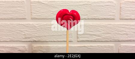 Lollipop in Form von rotem Herz gegen eine weiße Ziegelwand, Valentinstag Stockfoto