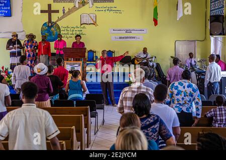 Ein Prediger in rotem Hemd und Krawatte singt mit ausgestreckten Armen vor der Bühne einer Baptistengemeinde in The Lime, Grenada, während eine Band im Hintergrund Gitarre und Schlagzeug spielt und mehrere Sängerinnen am Podium stehen. Ein Banner an der Wand kündigt „Ignited Generation Month“ im November an. Stockfoto