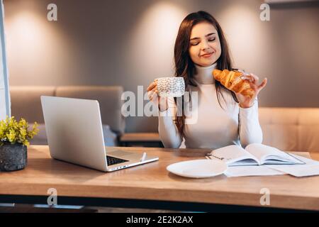 Freiberufliche Frau mit Laptop, während zu Hause sitzen. Junge Frau sitzt in der Küche und arbeitet am Laptop. Hübsche Frau trinkt Tee Kaffee während der Arbeit Stockfoto