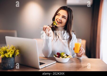 Fröhliche junge Frau trinkt Orangensaft und genießt es, Salat aus einer Schüssel zu essen, während sie auf einer Küche steht und Film auf dem Laptop sieht. Stockfoto