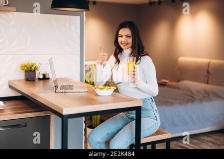 Junge lächelnde Frau beim Frühstück in der Küche, sie verbindet sich mit einem Laptop und trinkt einen gesunden Orangensaft. Stockfoto