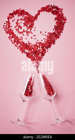 Zwei Sektgläser mit roten Bonbons in Herzform, groß Rotes Herz aus Süßigkeiten als Spritzer Champagner auf Rosa Hintergrund.Konzept für Begrüßung ca. Stockfoto