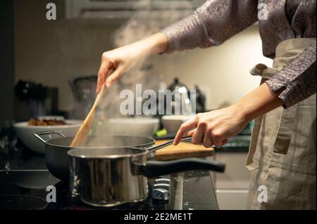 Frau in Schürze Kochen auf Keramik-Kochfeld in modernen Hause, Nahaufnahme der Hände Rührtopf in der Küche. London, Großbritannien Stockfoto
