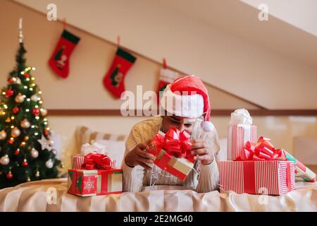 Gemischter Mann mit weihnachtsmann Hut auf dem Kopf liegend auf dem Bett und versucht, Weihnachtsgeschenk zu öffnen. Um ihn herum Geschenke, im Hintergrund Weihnachtsschmuck. Stockfoto