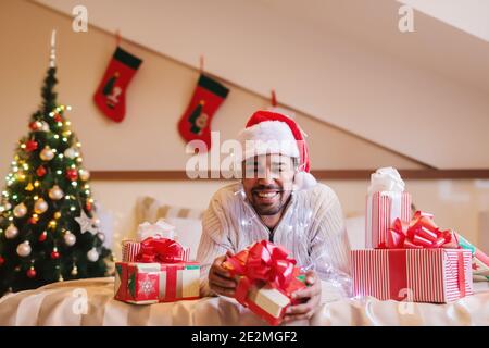 Lächelnder gemischter Rennmensch mit weihnachtsmann-Hut auf dem Kopf posiert auf dem Bett mit Geschenk in den Händen. Auf dem Bett Geschenke, im Hintergrund Weihnachtsbaum und Dekorationen. Stockfoto