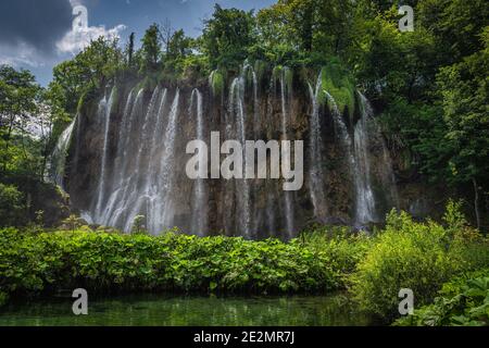 Streifen von Wasser aus Wasserfall durch Sonnenlicht beleuchtet. Kleiner Teich im grünen üppigen Wald. Nationalpark Plitvicer Seen UNESCO Weltkulturerbe, Kroatien Stockfoto