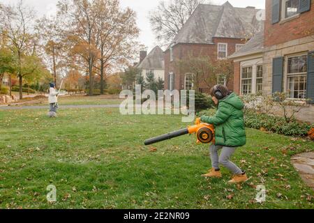 Ein Junge räumt Blätter von Rasen mit Laubbbläser während Mädchen Rechen Stockfoto