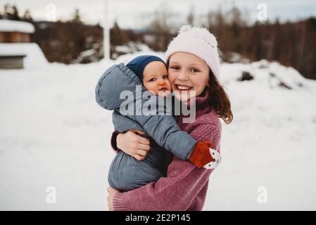 Smiley große Schwester hält Baby Bruder draußen im Schnee Bei kaltem da Stockfoto