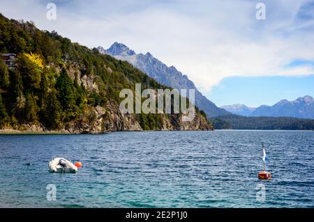 Seeufer mit Felsen, Bergen und Pinien. Ein Motorboot, das in der Nähe einer Boje schwimmt. Sommer in Bariloche. Stockfoto