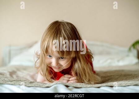 Lächelndes Baby auf dem Bett sitzend.nettes rothaariges Mädchen spielt zu Hause in einem hellen Schlafzimmer. Lustig und verspielt. In einem roten Kleid. Glückliche Kindheit Stockfoto