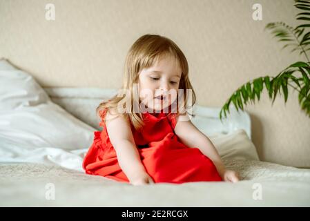 Lächelndes Baby auf dem Bett sitzend.nettes rothaariges Mädchen spielt zu Hause in einem hellen Schlafzimmer. Lustig und verspielt. In einem roten Kleid. Glückliche Kindheit Stockfoto
