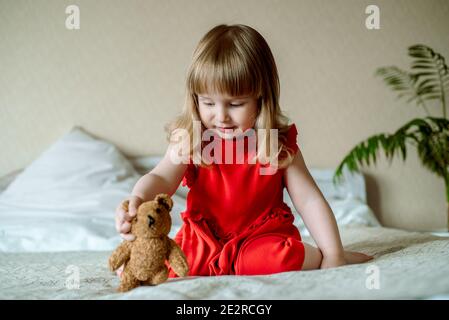 Lächelndes Baby auf dem Bett sitzend.nettes rothaariges Mädchen spielt zu Hause in einem hellen Schlafzimmer. Lustig und verspielt. In einem roten Kleid. Glückliche Kindheit.Teddybär. Stockfoto