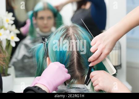 Rückansicht des weiblichen Kopfes mit smaragdgrüner Haarfarbe und nachwachsende Haarwurzeln. Frau sitzt in Stuhl durch Spiegel, zwei professionelle Friseure kämmen Kunden Stockfoto