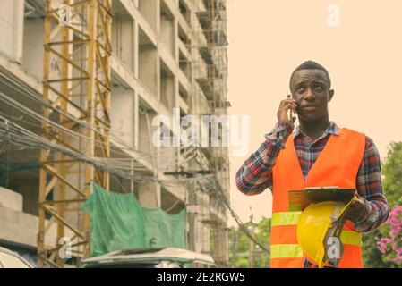 Nachdenklichen jungen schwarzen afrikanischen Mann Bauarbeiter Gespräch am Handy halten Helm und Zwischenablage an Baustelle Stockfoto