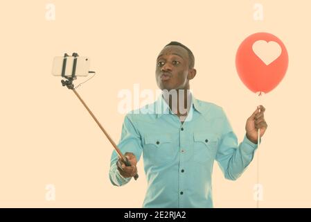Studio shot von jungen schwarzen afrikanischen Mann mit roten Ballons mit Herz Zeichen während verziehenden Lippen und unter selfie Bild mit Handy auf selfie Stockfoto