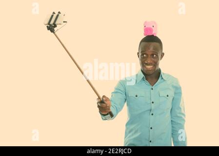 Studio shot der jungen schwarzen afrikanischen Mann glücklich lächelnd mit Sparschwein auf dem Kopf während der Einnahme von selfie Bild mit Handy auf selfie Stick Stockfoto