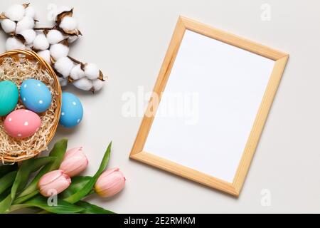 Frohe Ostern. Bunte Eier in einem Korb, Tulpen, ein Baumwollzweig und ein Rahmen auf einem grauen Hintergrund. Platz zum Kopieren von Grußkarten mit flachem Lay-Papier. Stockfoto