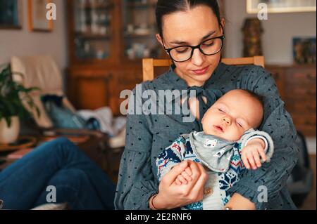 Junge Mutter sitzt im Wohnzimmer und hält neugeborenes Baby