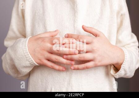 Nahaufnahme der Hände eines Kindes, die mit verflochtenen Fingern auf dem Bauch gefaltet sind, in einem hellen gestrickten champagnerfarbenen Pullover Stockfoto