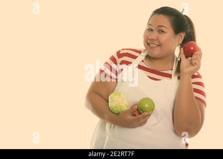 Studio shot der Jungen gerne Fett asiatische Frau lächelnd, während Sie red apple mit Obst und Gemüse beim Tragen Schürze Stockfoto