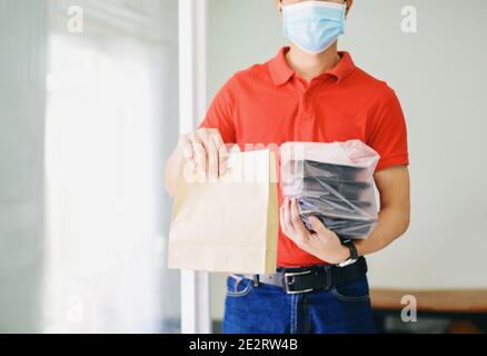 Lieferung Mann trägt schützende Gesichtsmaske Coronavirus und Hand halten Handwerk Papier Lebensmittelverpackung Service, Postmann und drücken verschiedene Papier contai Stockfoto